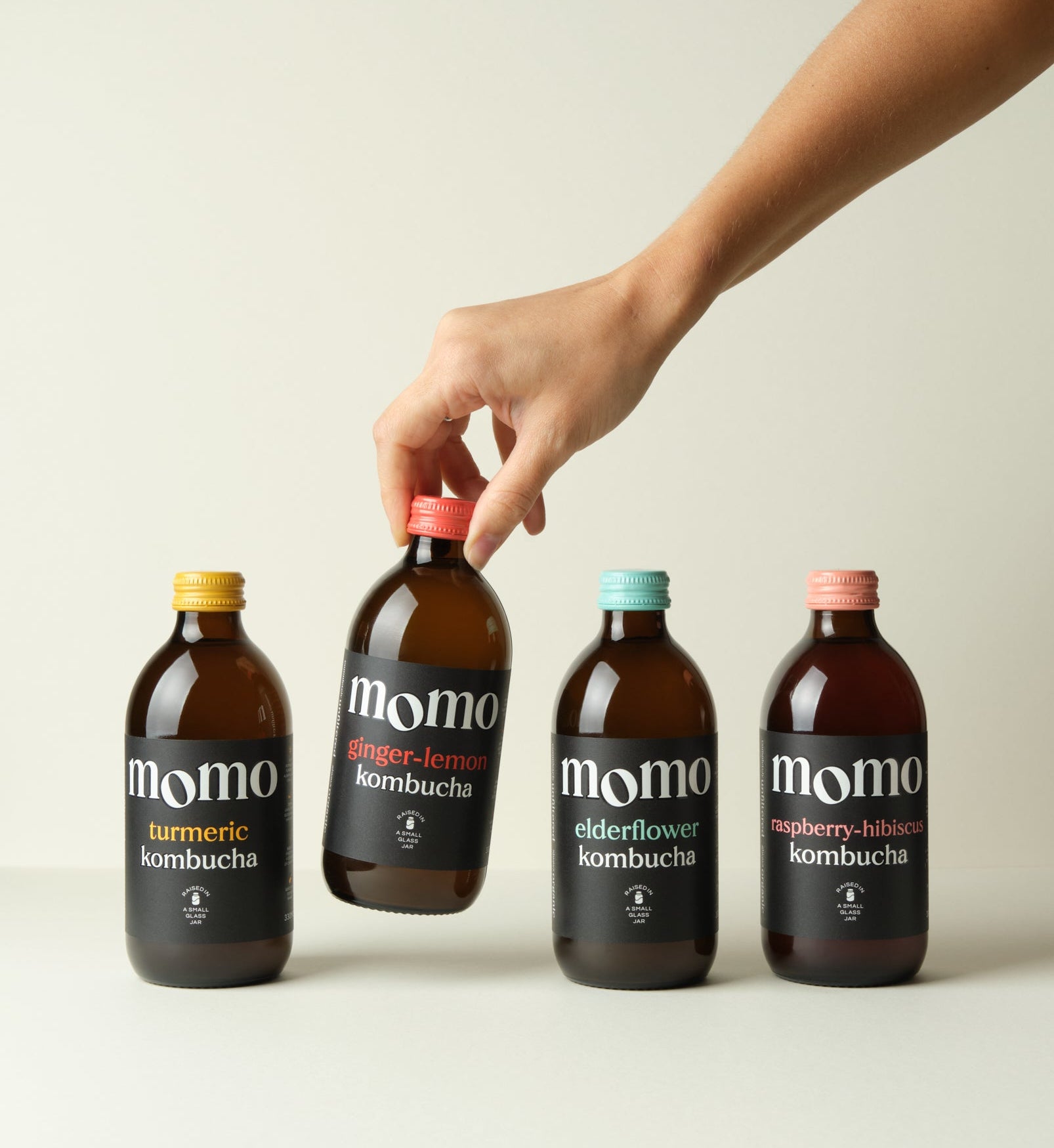 Hand picking a bottle of momo kombucha from a set of four on a light background